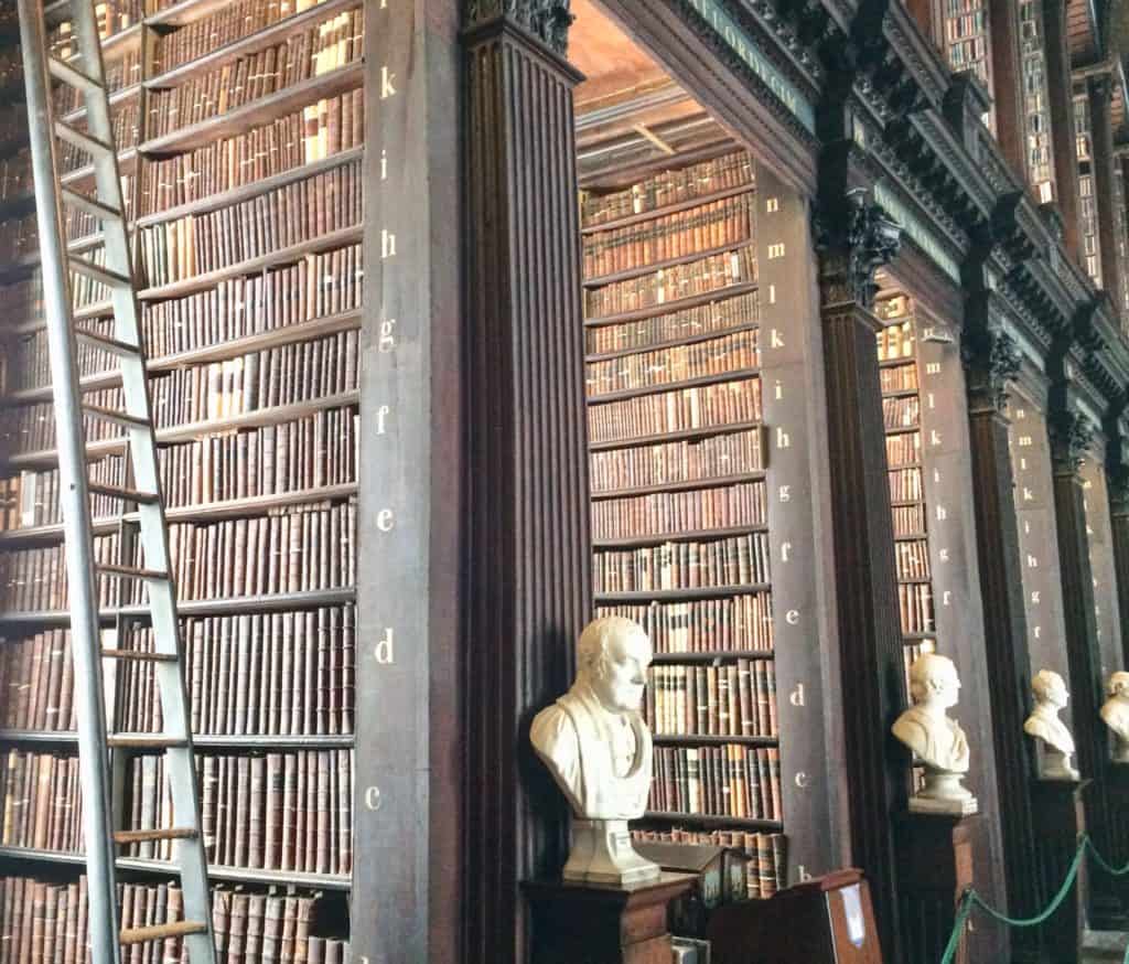 Ladder Near Rows Of Tall Bookshelves With Old Books and Marble Busts Receding Into Distance