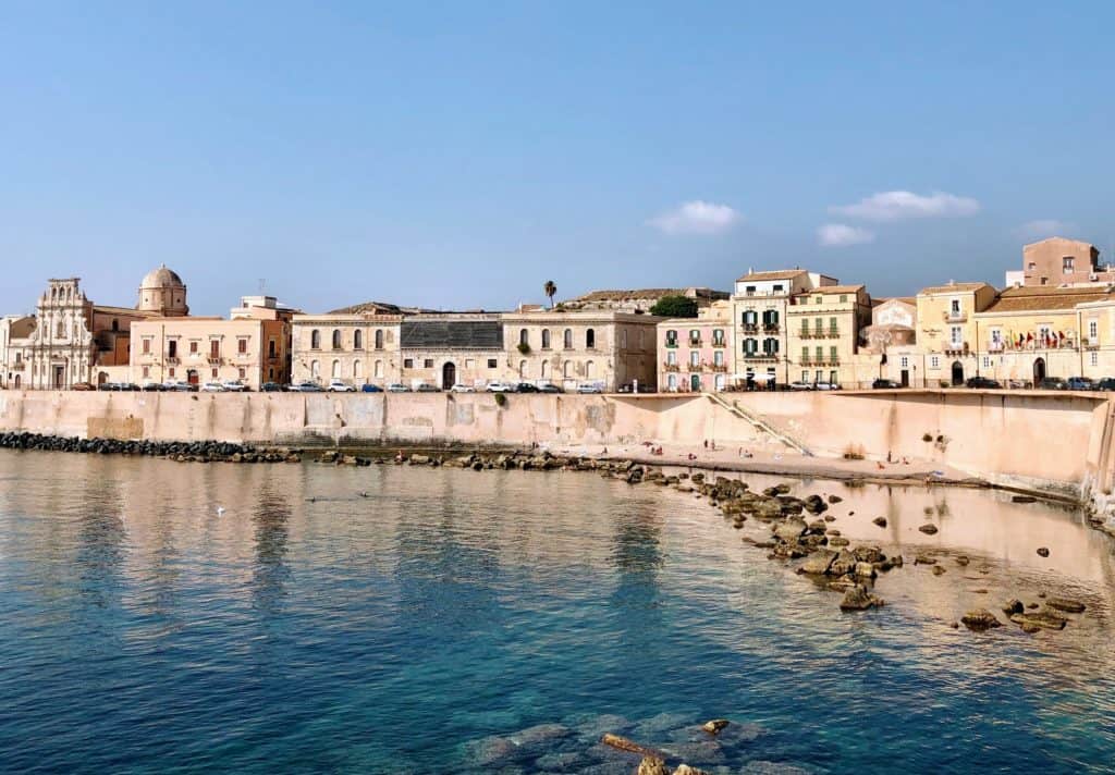 Syracuse Italy Curving Wall Between Weathered Pastel Buildings And Sea With Rocks